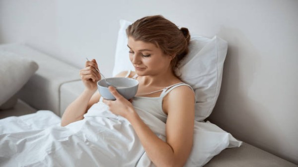 Ilustrasi seseorang menikmati sup hangat di tempat tidur saat flu, ditemani teh dan perlengkapan obat.