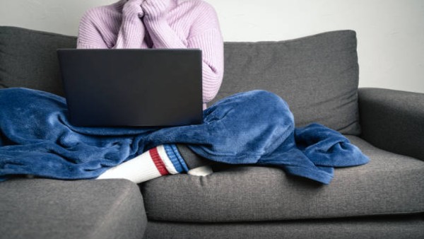 Ilustrasi seseorang mengenakan sweater dan selimut saat bekerja di sofa karena merasa kedinginan.