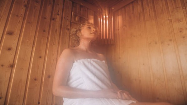 Seorang wanita sedang bersantai di ruang sauna kayu modern dengan uap panas yang memenuhi ruangan, menggambarkan relaksasi sekaligus manfaat kesehatan dari terapi panas.